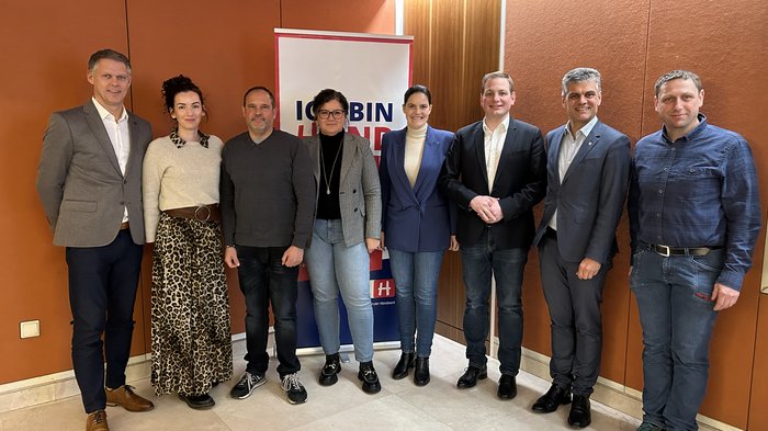 Arbeit durch Bürokratie erschwert Acht Personen stehen zusammen vor einer Werbetafel mit orangefarbenem Hintergrund