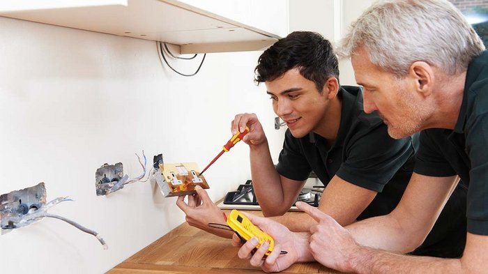Lohnausgleichskasse fängt auch Lehrlinge im Bausektor auf Zwei Elektriker installieren eine Steckdose in einer Küche