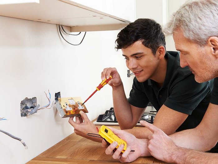 Lohnausgleichskasse fängt auch Lehrlinge im Bausektor auf Zwei Elektriker installieren eine Steckdose in einer Küche