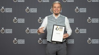 News Mann hält German Design Award 2025 Urkunde, lächelnd vor Award-Hintergrund
