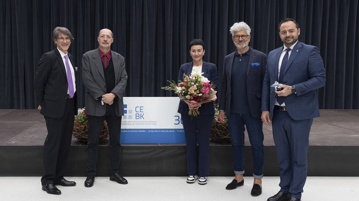 Cassa Edile della Provincia Autonoma di Bolzano: premiazione iscritti “30 anni di fiducia“ Cinque persone posano davanti a una tenda, una tiene un mazzo di fiori, una un regalo.