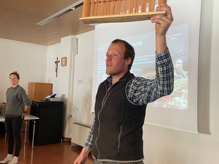 Junghandwerker/innen im lvh und Hoteliers- und Gastwirtejugend (HGJ) zu Gast in der Mittelschule Sarntal Mann hält ein Holzmodell in einem Vortragsraum hoch