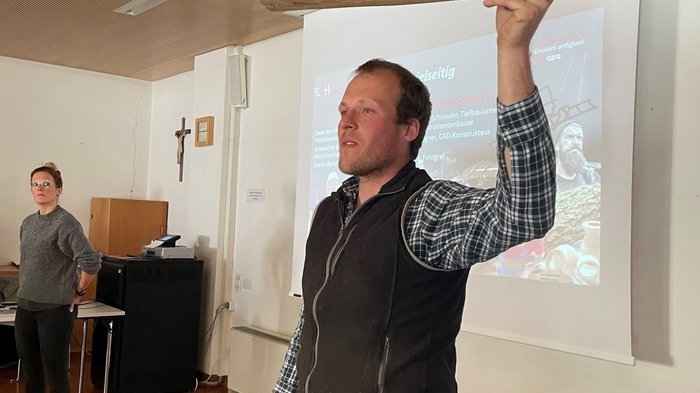 Junghandwerker/innen im lvh und Hoteliers- und Gastwirtejugend (HGJ) zu Gast in der Mittelschule Sarntal Mann hält ein Holzmodell in einem Vortragsraum hoch