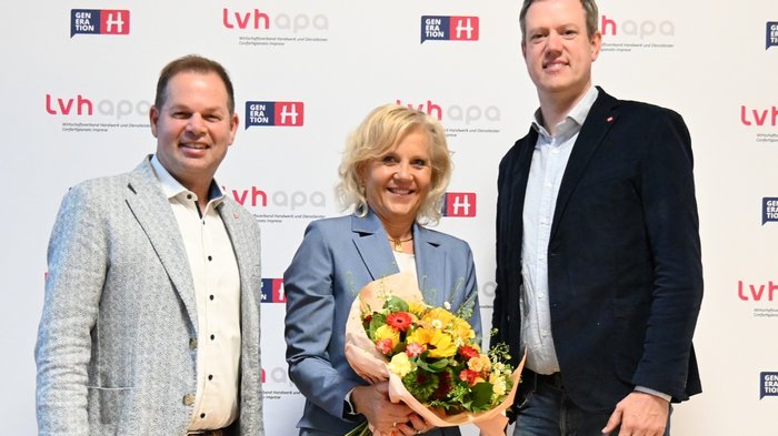 Versicherungen für Handwerksbetriebe im Überblick Drei Personen lächeln, eine mit Blumenstrauß vor lvhapa-Hintergrund