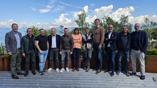 News Zwölf Personen stehen draußen auf Holzterrasse mit Bäumen und Bergen im Hintergrund