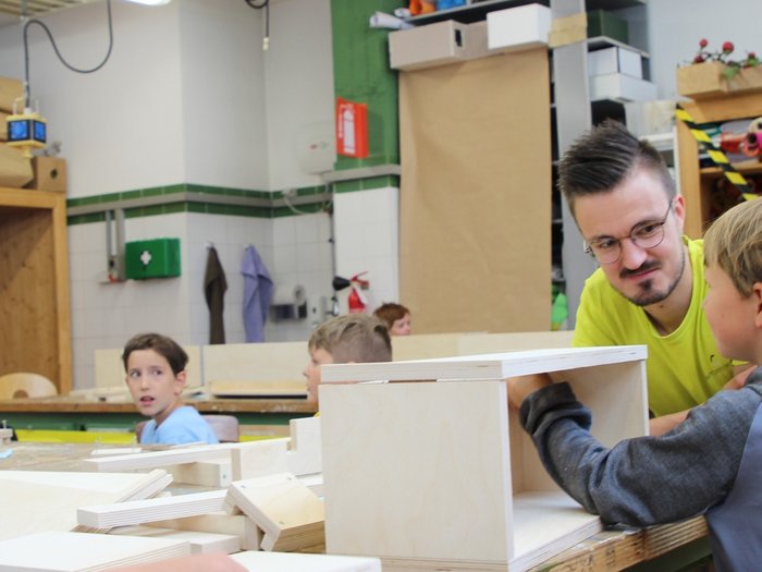 Kids-Academy 2024: Ein Sommer voller Kreativität und Handwerk Lehrer unterstützt Kinder beim Holzarbeiten in einer Werkstatt