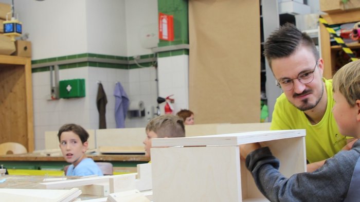 Kids-Academy 2024: Ein Sommer voller Kreativität und Handwerk Lehrer unterstützt Kinder beim Holzarbeiten in einer Werkstatt