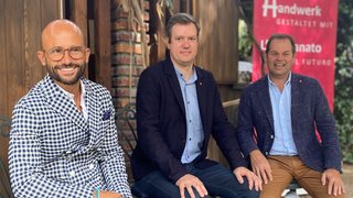 News Drei Männer sitzen vor Holzhaus mit Banner zum Thema Handwerk und Zukunft
