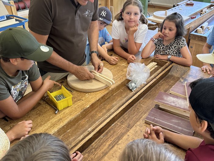 Kids Academy 2024: a Brunico l’artigianato colora l’estate dei più piccoli Insegnante mostra ai bambini lavori in legno in laboratorio