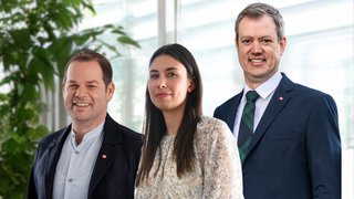 News Drei lächelnde Personen stehen vor einem Fenster mit Pflanzen im Hintergrund