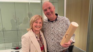 News Zwei lächelnde Personen in einem Büro, eine hält einen Block, der andere eine Rolle