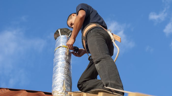 Holzofen: Im Winter auf Nummer sicher gehen Handwerker installiert isoliertes Rohr auf Dach bei klarem Himmel
