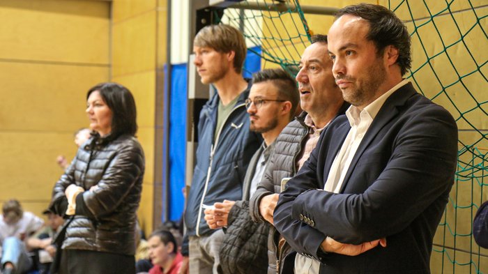 Kreative Spielideen: Preisverleihung für "Clever gebaut" Fünf Personen beobachten konzentriert ein Sportereignis in der Halle