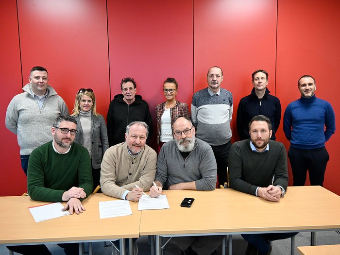 Un passo importante per l’artigianato edile altoatesino Gruppo di undici persone davanti a parete rossa, due uomini firmano documenti al tavolo