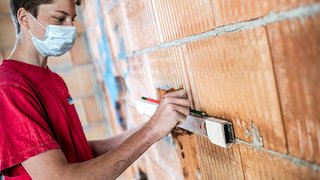 News Handwerker mit Maske misst und markiert Mauerwerkswand mit Wasserwaage und Bleistift