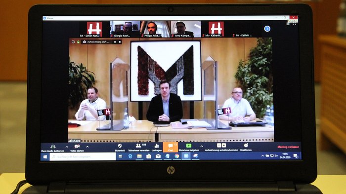 „Wieder arbeiten zu dürfen, ist die wichtigste Basis für den Neustart“ Laptopbildschirm zeigt Videokonferenz mit drei Personen an einem Tisch und mehreren Teilnehmern