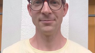 News Porträt eines Mannes mit Brille in gelbem T-Shirt vor weißer Wand