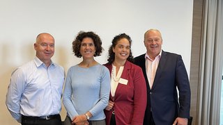 News Vier Personen stehen nebeneinander vor einer weißen Wand und lächeln in die Kamera