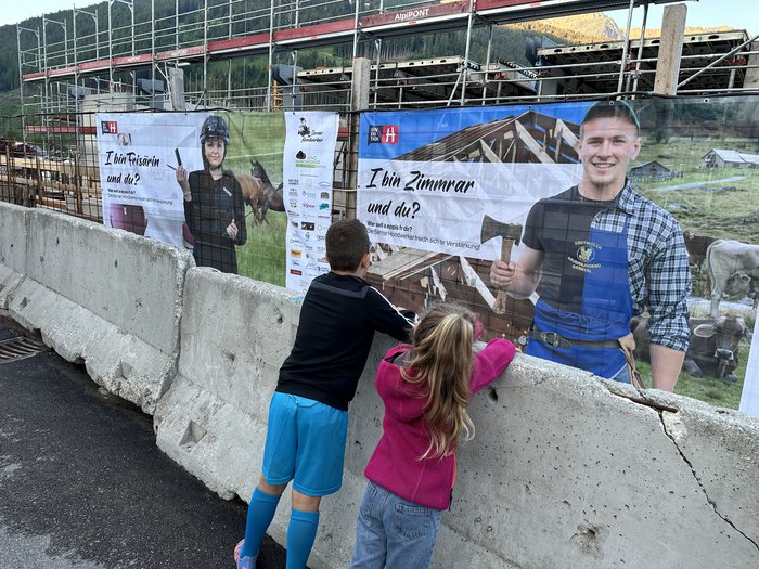 Jugend motivieren! Zwei Kinder schauen sich Bauplakate mit Handwerksberufen auf einer Baustelle an