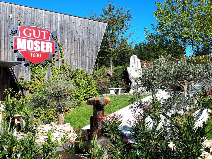 Unsere Althandwerker zu Besuch in der Stiermark Garten mit Brunnen und großer Hand-Skulptur neben Gebäude mit Gut Moser Schild