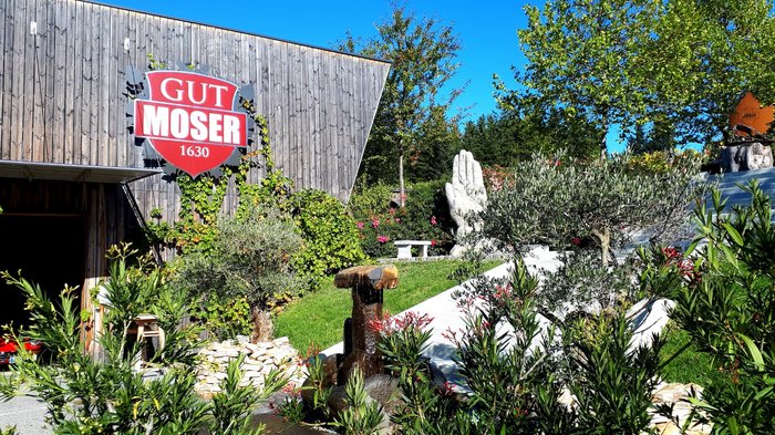 Unsere Althandwerker zu Besuch in der Stiermark Garten mit Brunnen und großer Hand-Skulptur neben Gebäude mit Gut Moser Schild