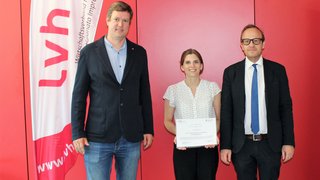 Foto - Uni Innsbruck © lvh.apa Drei Personen vor roter Wand, eine Frau hält Urkunde vor LVH-Fahne