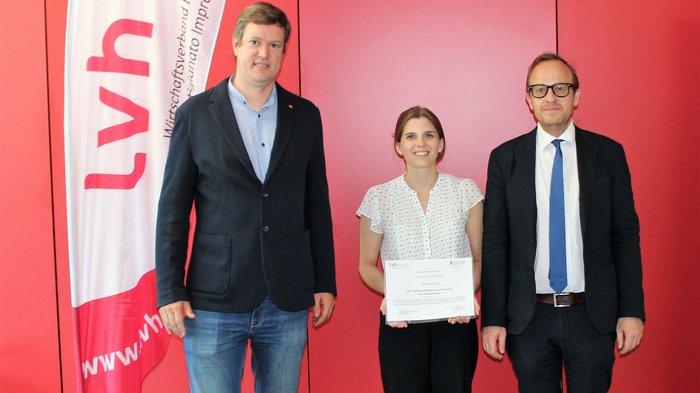 Foto - Uni Innsbruck © lvh.apa Drei Personen vor roter Wand, eine Frau hält Urkunde vor LVH-Fahne