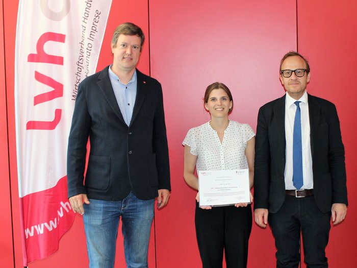 Foto - Uni Innsbruck © lvh.apa Drei Personen vor roter Wand, eine Frau hält Urkunde vor LVH-Fahne