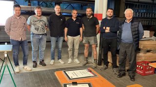 News Sieben Männer stehen in einer Werkstatt neben einem Tisch mit Urkunden