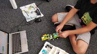 News Kind baut Roboterauto mit LEGO Education Set neben Laptop auf grauem Teppich
