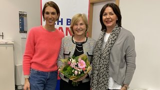 News Drei Frauen stehen zusammen, eine hält einen Blumenstrauß in einem Büro