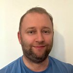 Unterland Porträt eines lächelnden Mannes mit kurzem Bart und blauem T-Shirt vor weißer Wand