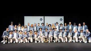 News Gruppe in blauen Jacken und Hüten auf einer Bühne vor hellen Leinwänden