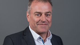 News Porträt eines lächelnden Mannes in schwarzem Anzug und weißem Hemd vor grauem Hintergrund