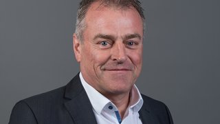 News Ritratto di uomo sorridente con giacca nera e camicia bianca su sfondo grigio