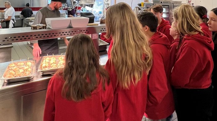 Eintauchen in die Welt der Berufe in Mühlbach Kinder in roten Pullovern stehen in einer Küche vor einem Mann und Tabletts mit Burgern