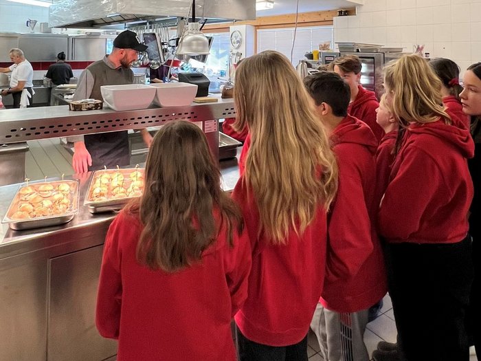 Eintauchen in die Welt der Berufe in Mühlbach Kinder in roten Pullovern stehen in einer Küche vor einem Mann und Tabletts mit Burgern