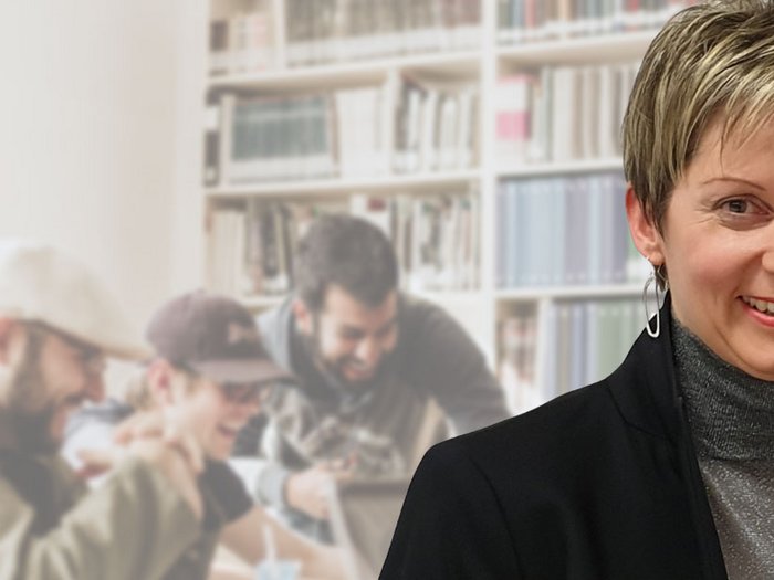 Staatliche Matura-Prüfung über den Berufsbildungsweg - Nun auch berufsbegleitend möglich! Frau lächelt vor verschwommenem Hintergrund mit Personen in Bibliothek