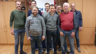 News Acht Männer unterschiedlichen Alters in einem Raum mit Holzmöbeln und Banner