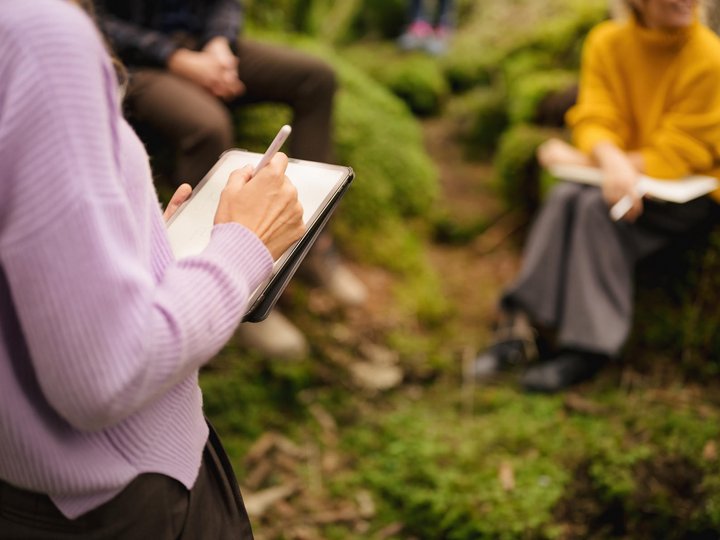 Das Nachhaltigkeitslabel Südtirol Person schreibt auf Tablet, andere sitzen im Wald auf Moos und Notizbüchern