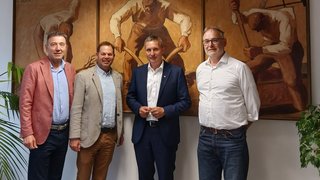 News Vier Männer stehen vor einem dreiteiligen Gemälde mit arbeitenden Figuren