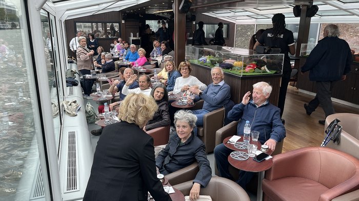 lvh-Seniorinnen und Senioren auf Frühlingsreise: Paris, mon amour! Gruppe älterer Menschen sitzt und unterhält sich in einem Bootscafé mit Panoramafenstern