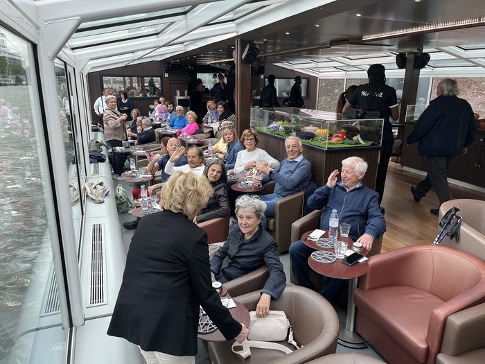 lvh-Seniorinnen und Senioren auf Frühlingsreise: Paris, mon amour! Gruppe älterer Menschen sitzt und unterhält sich in einem Bootscafé mit Panoramafenstern