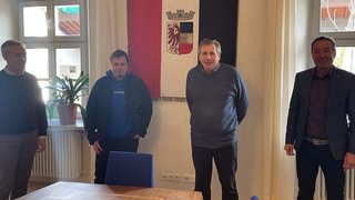 News Vier Männer stehen vor einer Wand mit einem Wappen in einem hellen Raum