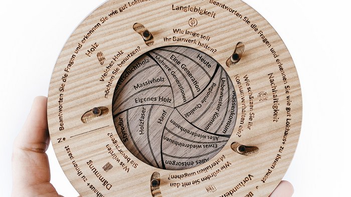 Gleich drei Auszeichnungen in Platin Hand hält runden, beschrifteten Holzdiagramm mit Fragen zu Holz und Nachhaltigkeit