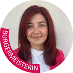 Burggrafenamt Porträt einer lächelnden Bürgermeisterin mit roten Haaren vor weißer Wand.