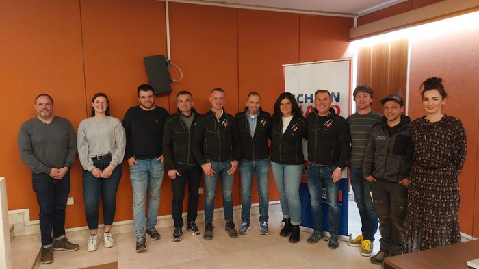 Lananer Handwerk: Frau an der Spitze Gruppe von elf Personen posiert vor oranger Wand in Innenraum