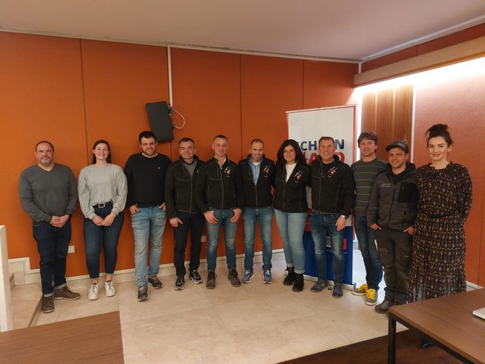 Lananer Handwerk: Frau an der Spitze Gruppe von elf Personen posiert vor oranger Wand in Innenraum