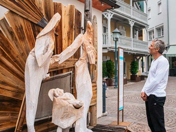 IDEA UNIKA 2025: 6ª edizione della Biennale d’arte nei centri dei paesi della Val Gardena Uomo osserva una natività in legno con figure in una strada cittadina