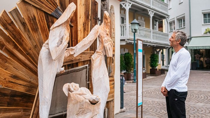 IDEA UNIKA 2025: 6ª edizione della Biennale d’arte nei centri dei paesi della Val Gardena Uomo osserva una natività in legno con figure in una strada cittadina